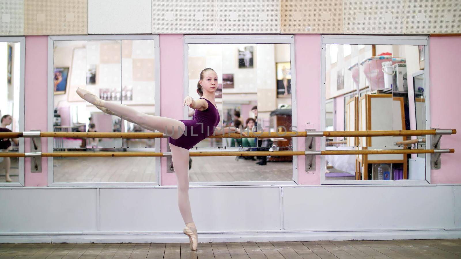 in dancing hall, Young ballerina in purple leotard performs developpe attitude on pointe shoes, raises her leg up behind elegantly, standing near barre at mirror in ballet class. High quality photo