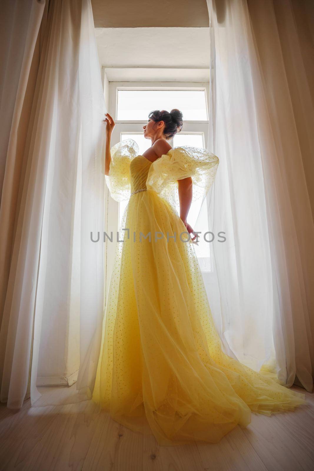 A woman's silhouette in a golden luxurious dress against the background of a window holds a curtain with her hands. Elegant lady in a yellow long silk dress with bare back, back view. by Matiunina