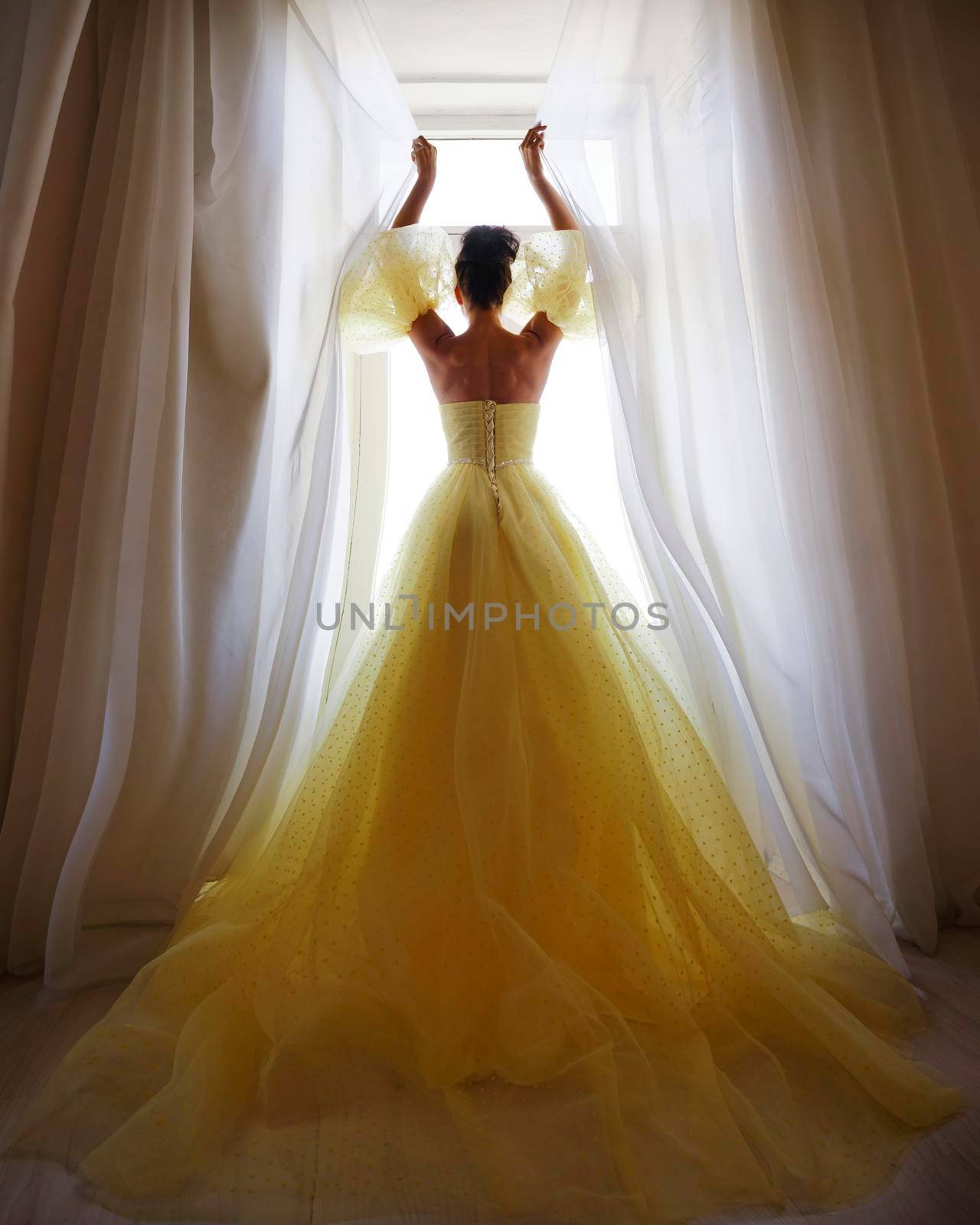 A woman's silhouette in a golden luxurious dress against the background of a window holds a curtain with her hands. Elegant lady in a yellow long silk dress with bare back, back view. by Matiunina