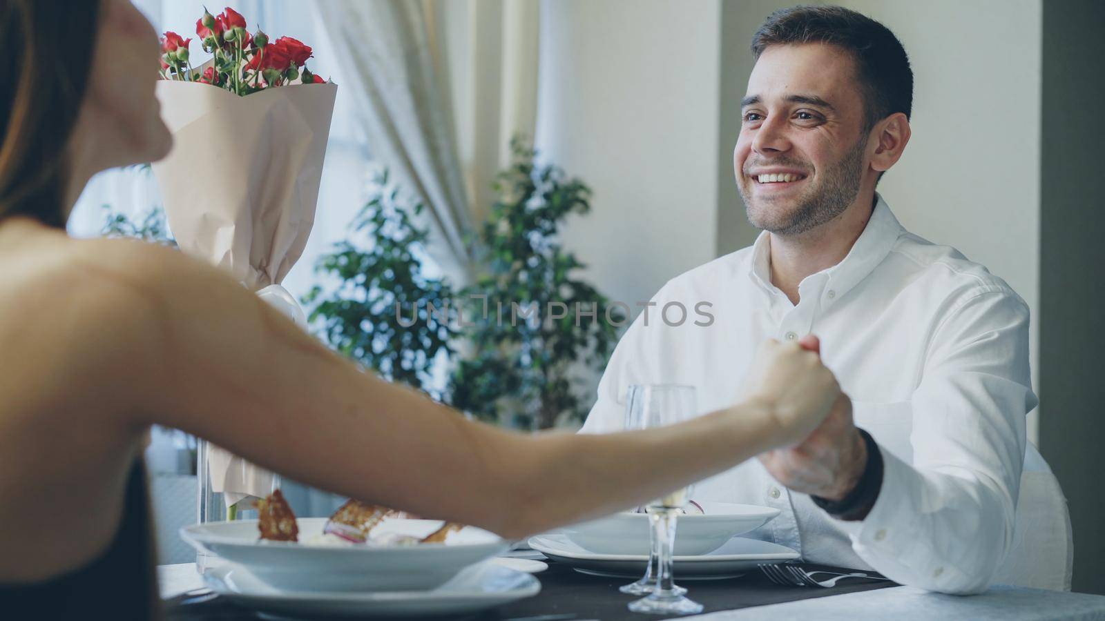 Young handsome man is talking to his girlfriend on date in fancy restaurant, drinking champagne and taking her hand with love and care. Romance and relationships concept.