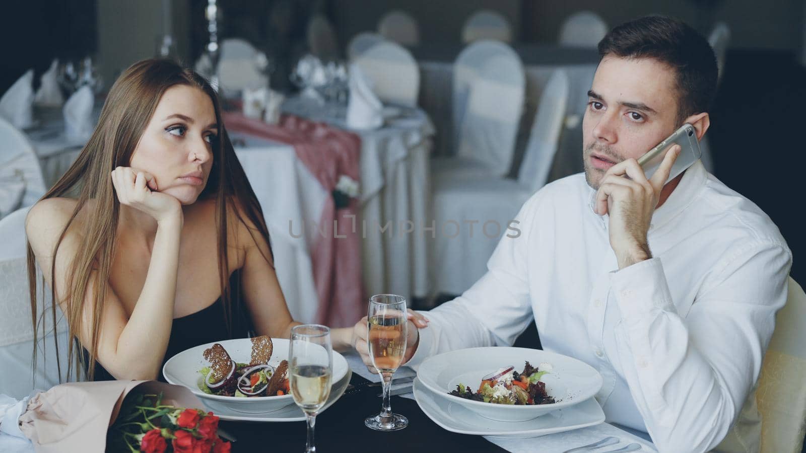 Busy young man is talking on mobile phone while his girlfriend is feeling bored sitting at table in restaurant. Modern technologies, relationship and boredom concept. by silverkblack