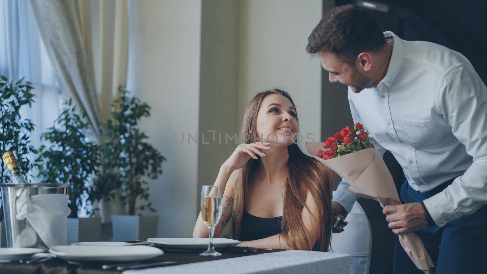 Happy girlfriend is getting flowers and present from her boyfriend after waiting for him alone in restaurant. Romantic relationship, gifts and fine dining concept. by silverkblack