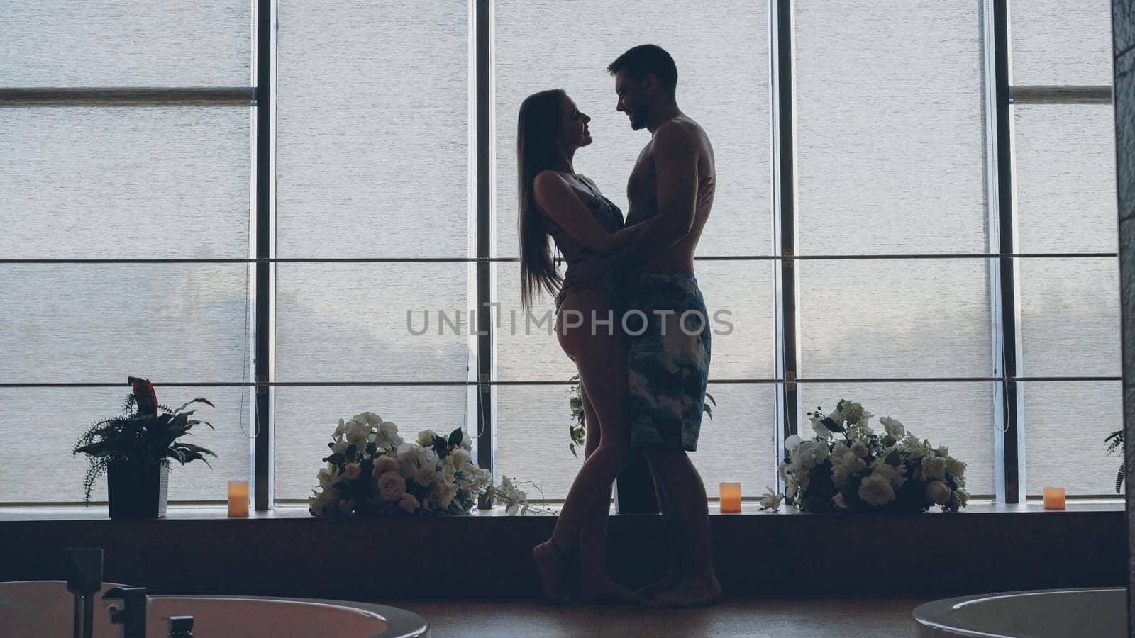 Silhouette of loving young people in bathing suits meeting in modern day spa and hugging . Candles, beautiful flowers, jacuzzi are visible.