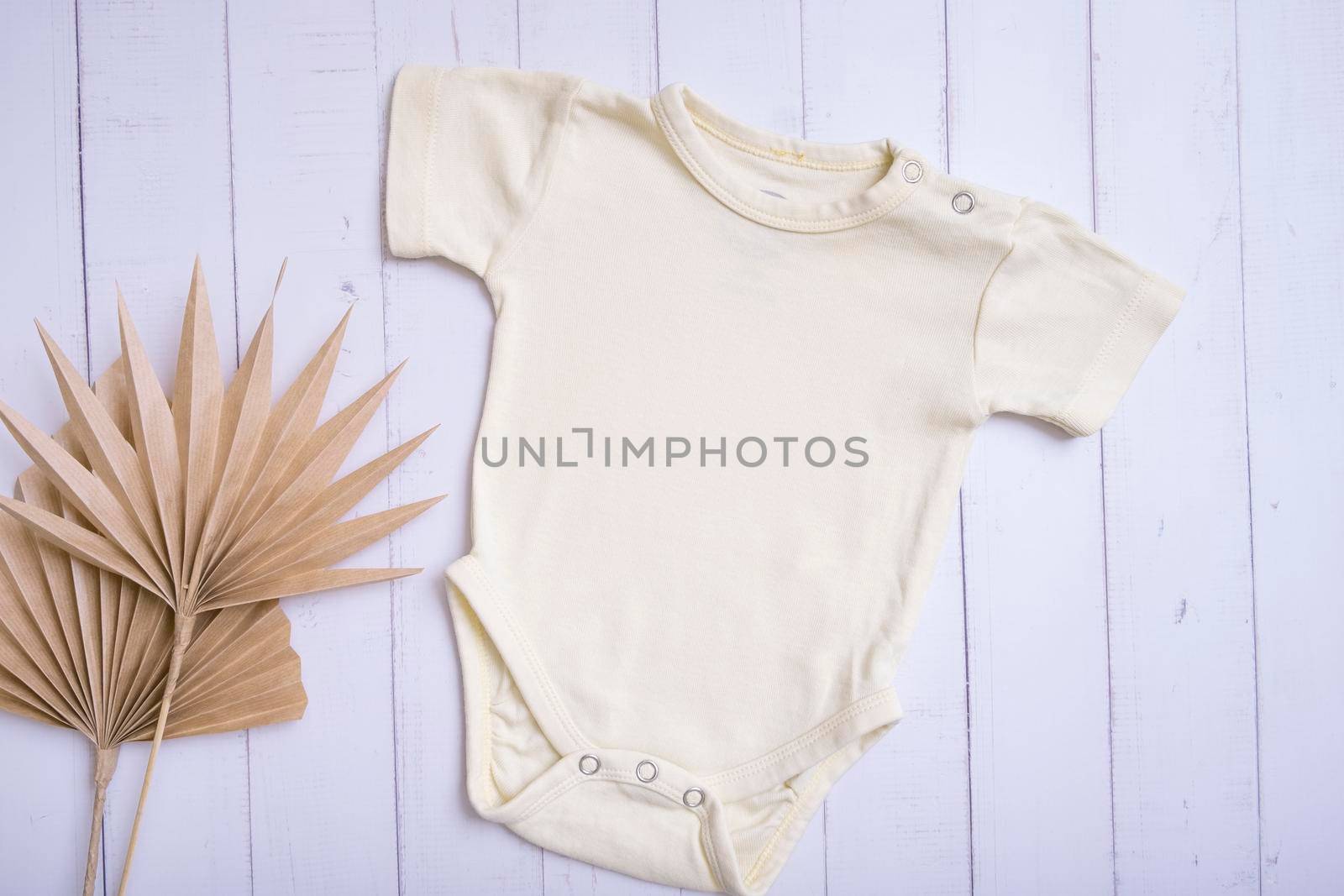 Yellow baby bodysuit mockup for logo, text or design on wooden background with palm leaves top view.