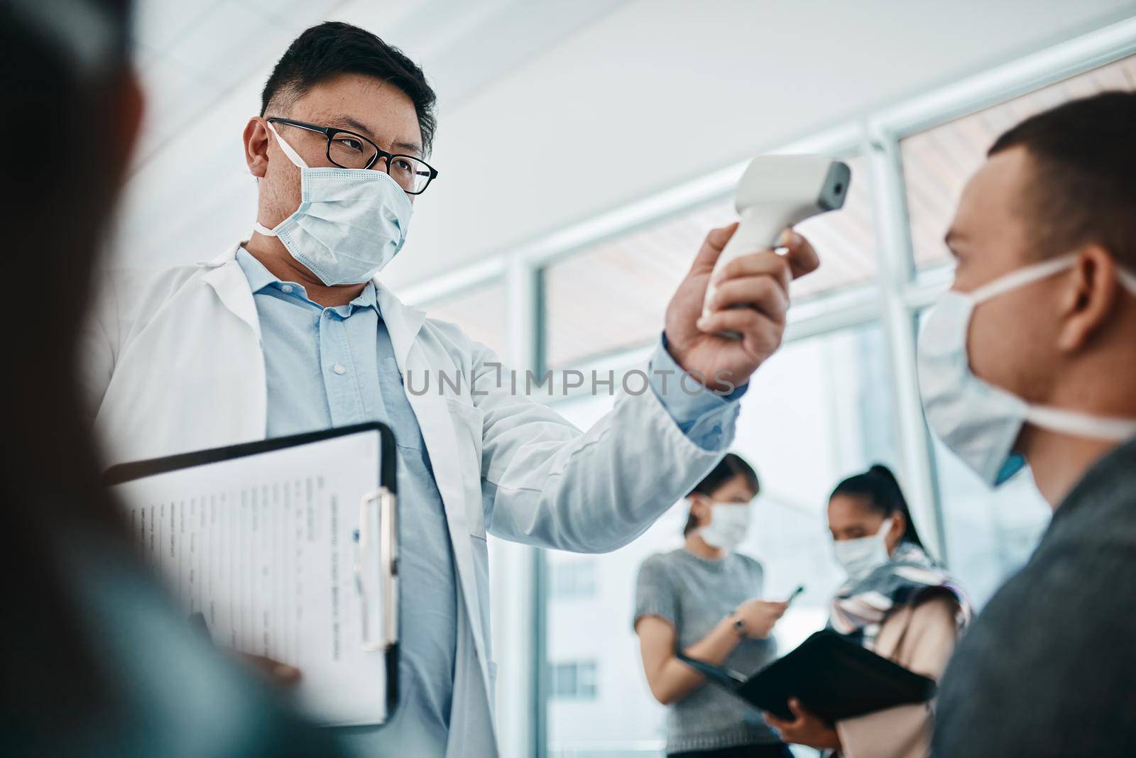 Doctor testing covid temperate in hospital appointment to prevent the spread of the virus. Healthcare compliance worker with health insurance document and face mask checking or scanning patient head by YuriArcurs
