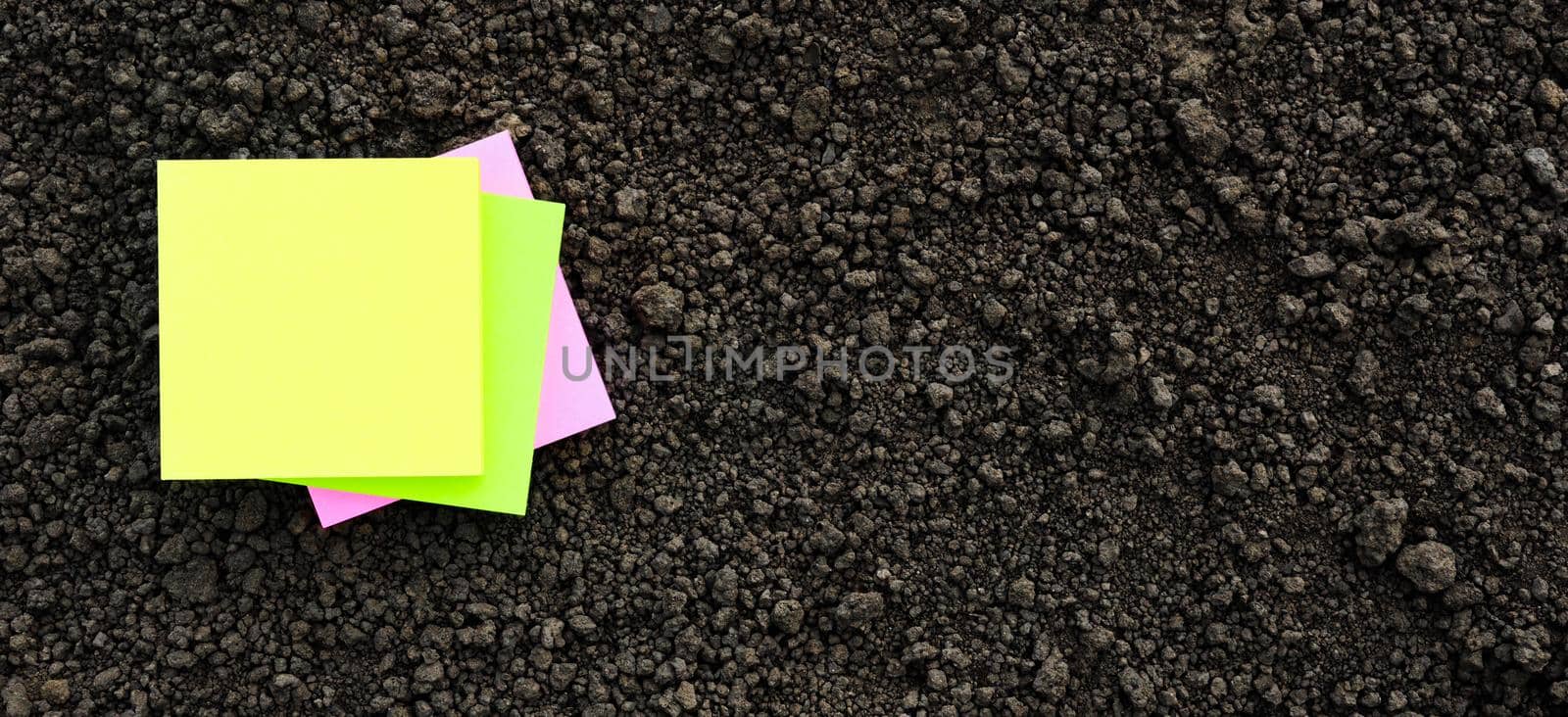 Yellow sticky note blank paper flat soil organic earth texture. Colors note paper square on black soil texture ground close up. Mockup on ground soil background top view from above. Agriculture banner by synel