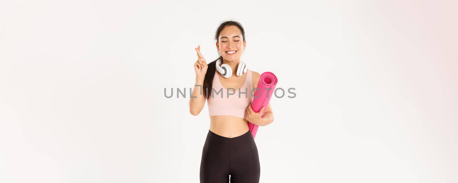 Sport, wellbeing and active lifestyle concept. Hopeful cute asian girl dreaming of going to gym after covid-19 quarantine, holding rubber mat and cross fingers while making wish, pleading by Benzoix