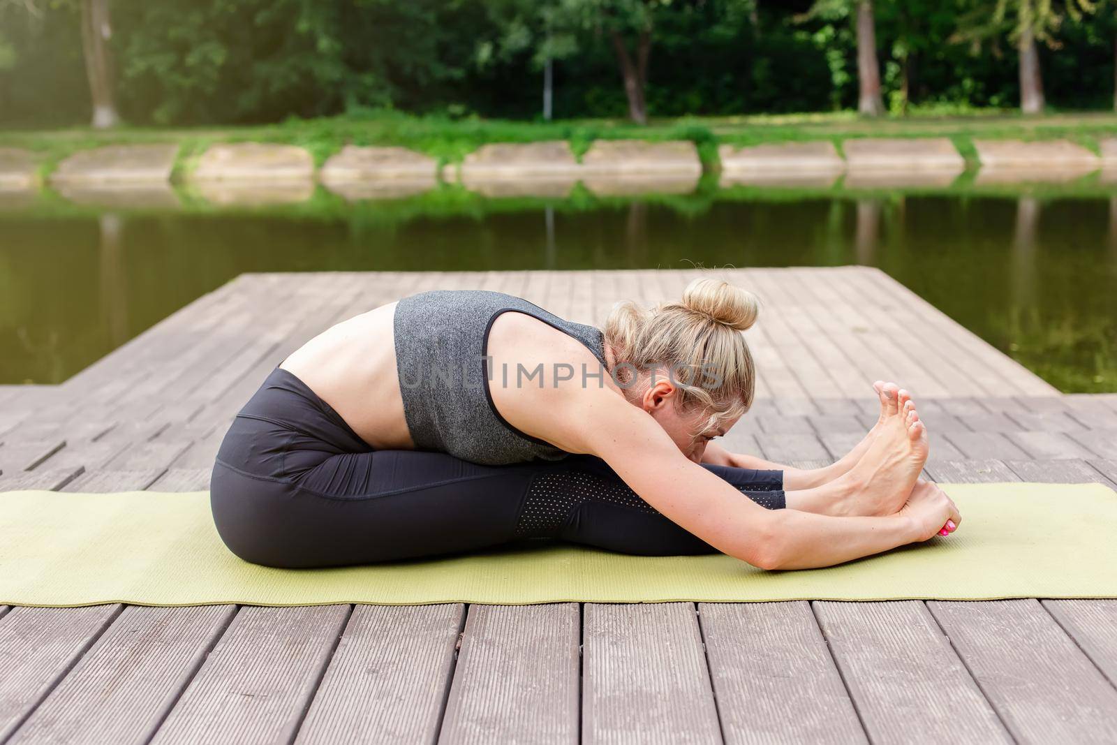 A woman in a gray top and leggings in summer on a wooden platform by a pond in the park does yoga, doing stretching. Copy space