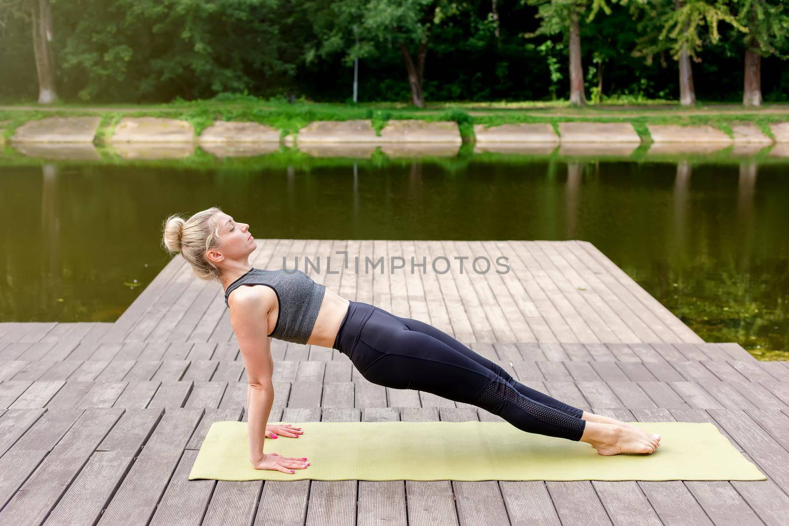 A woman in a gray top and leggings, on a wooden platform by a pond in the park in summer, does yoga, on a green mat, performing a reverse plank. Copy space