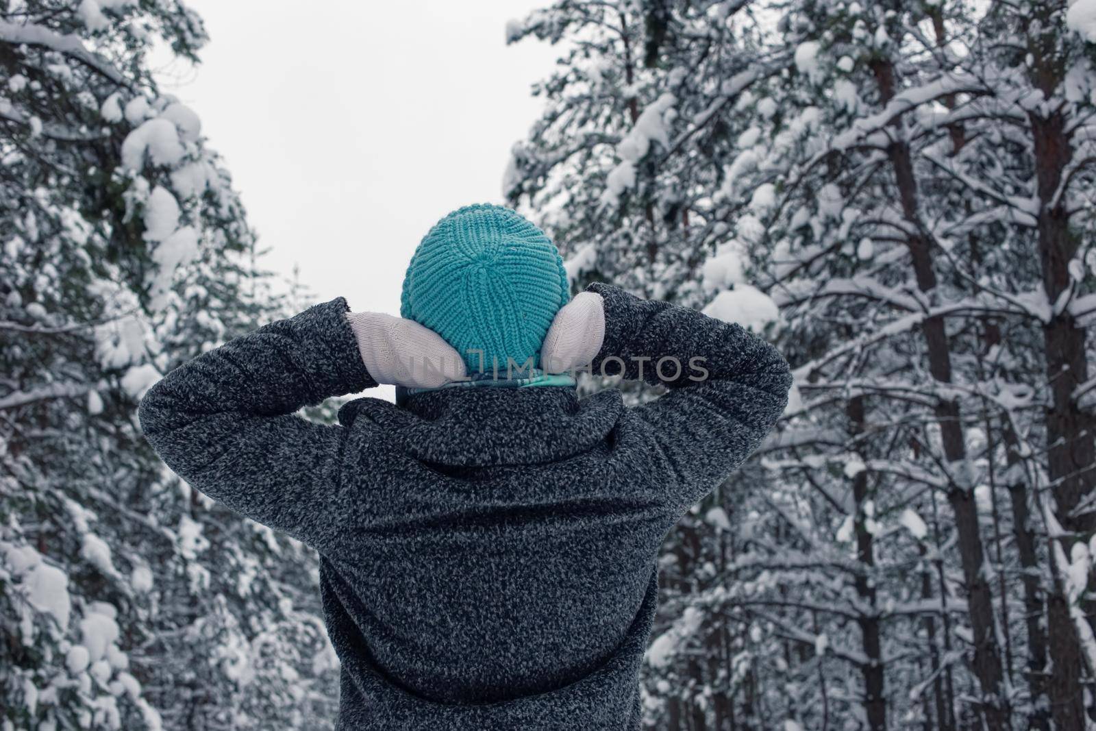 A girl in a gray coat and a light blue knitted hat stands in a beautiful winter snowy forest on a cloudy snowy day. Back view