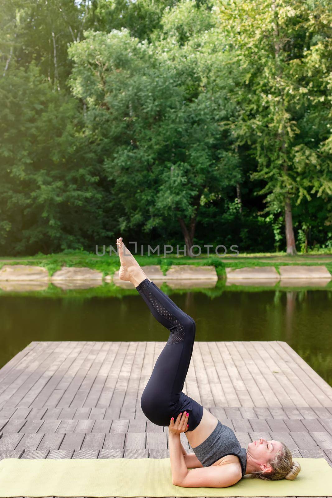 A slim woman in a gray top and leggings, on a wooden platform by a pond in a park in summer, does yoga, on green mat. Copy space. Vertical
