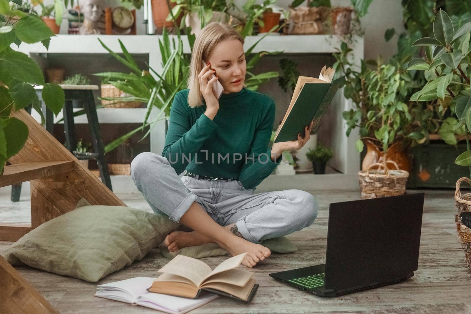 A blonde woman in a room with a lot of green indoor plants is working on a laptop. The concept of biophysical design in the interior. Work from home, work as a freelancer.