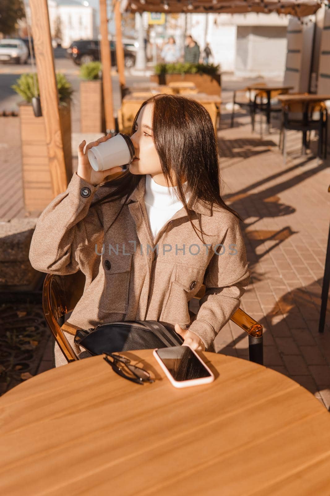 A stylish brunette woman is sitting at a table in an outdoor cafe with coffee and a phone. Autumn walk.