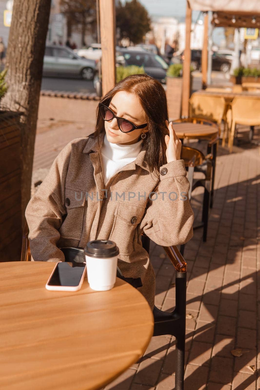 A stylish brunette woman in sunglasses is sitting at a table in an outdoor cafe with coffee and a phone. The girl is talking on the phone. Autumn walk.