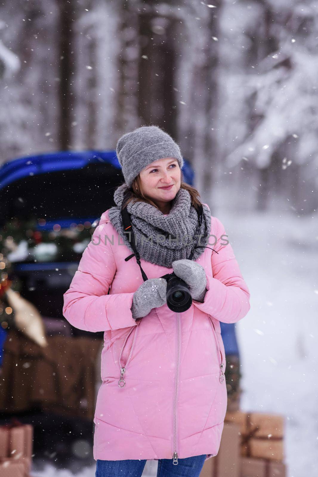 A woman in a winter snow-covered forest in the trunk of a car decorated with Christmas decor. A female photographer holds a camera in her hands.