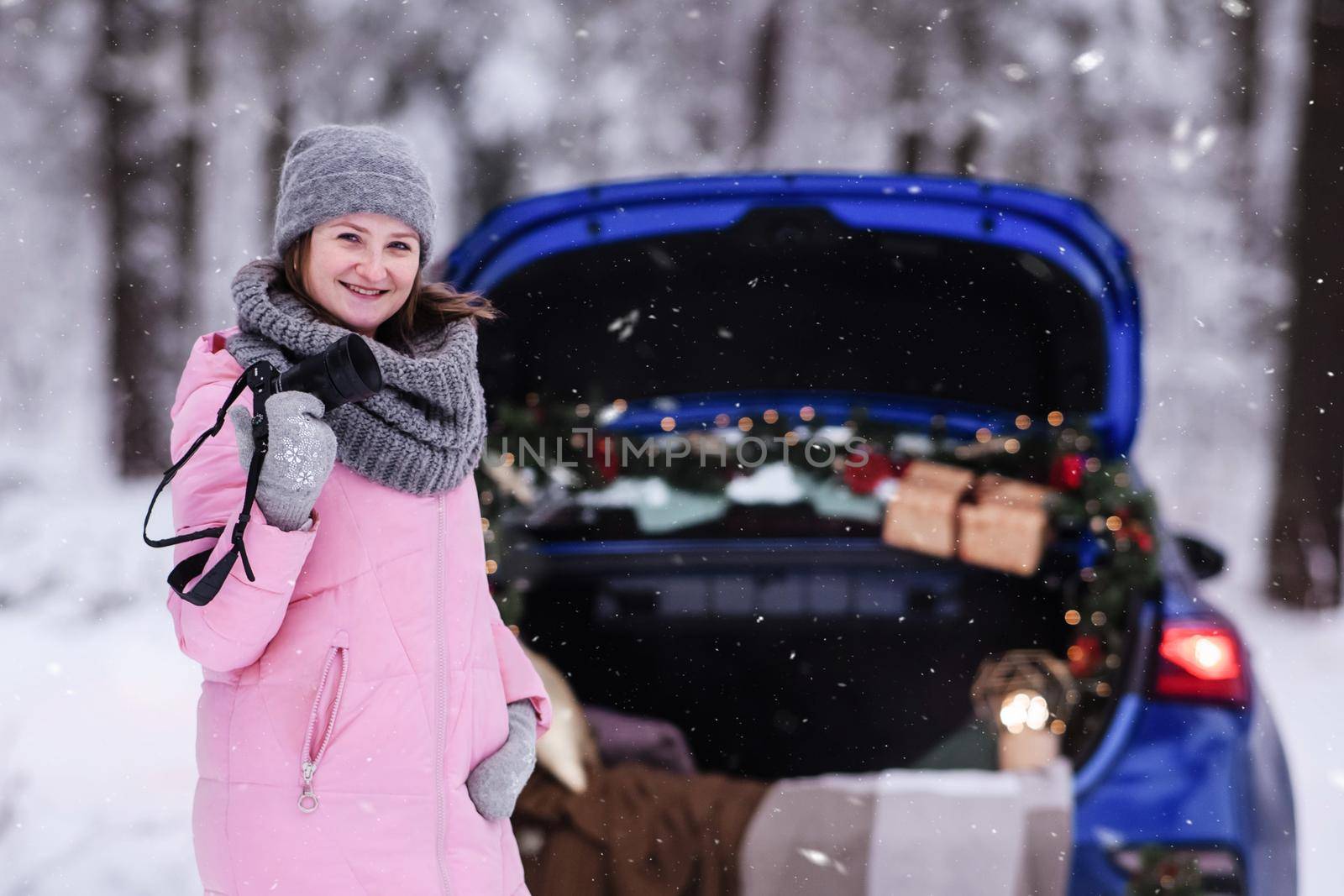 A woman in a winter snow-covered forest in the trunk of a car decorated with Christmas decor. A female photographer holds a camera in her hands.