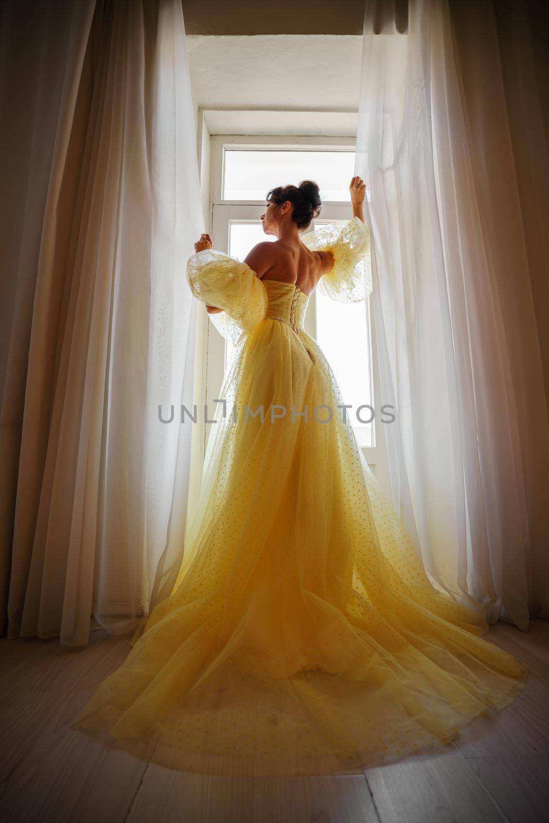 A woman's silhouette in a golden luxurious dress against the background of a window holds a curtain with her hands. Elegant lady in a yellow long silk dress with bare back, back view. by Matiunina