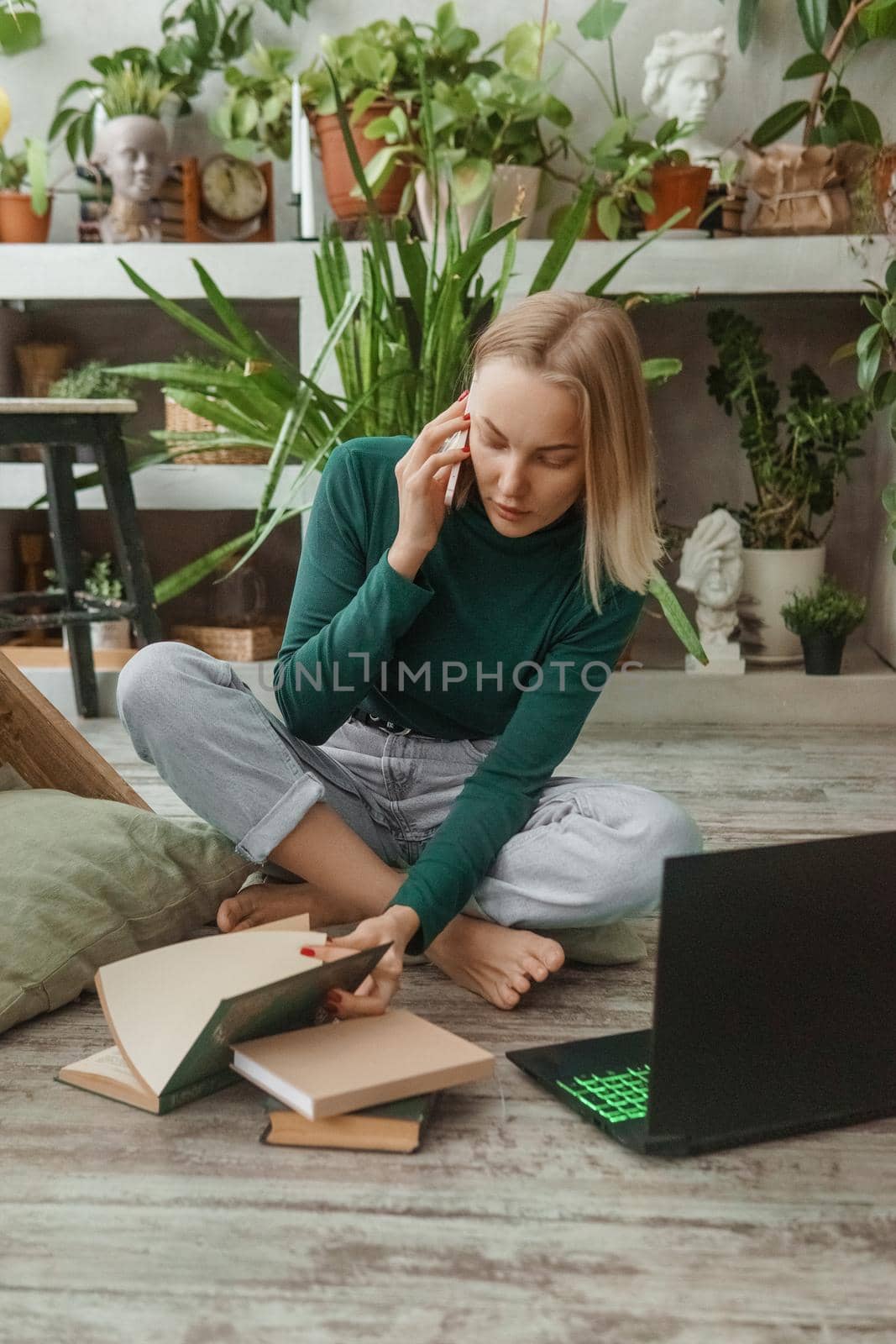 A blonde woman in a room with a lot of green indoor plants is working on a laptop. The concept of biophysical design in the interior. Work from home, work as a freelancer.