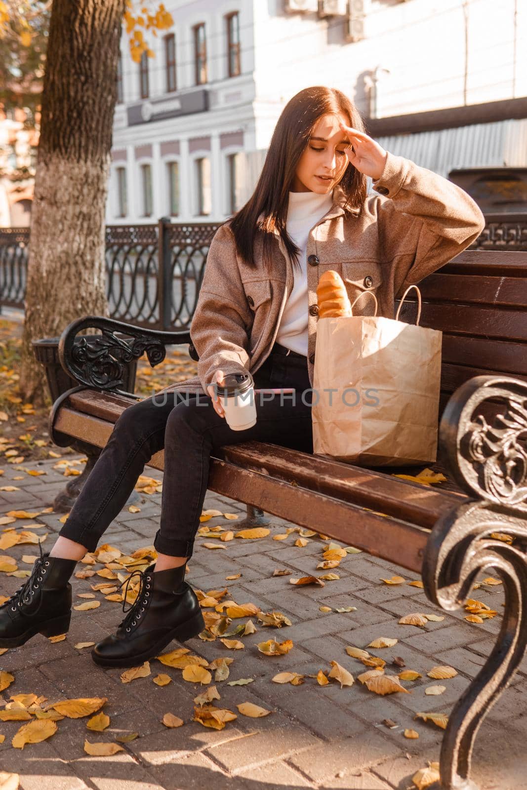 A stylish brunette woman walks around the autumn city. The brunette is sitting on a bench outside with a glass of coffee and a phone, holding a craft bag with a baguette.