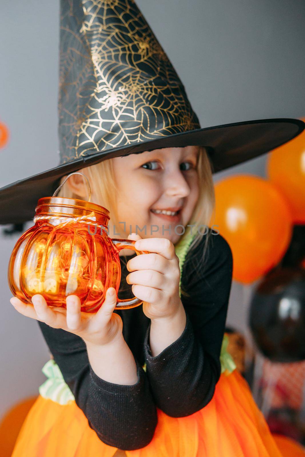 Children's Halloween - a girl in a witch hat and a carnival costume with airy orange and black balloons at home. Ready to celebrate Halloween.