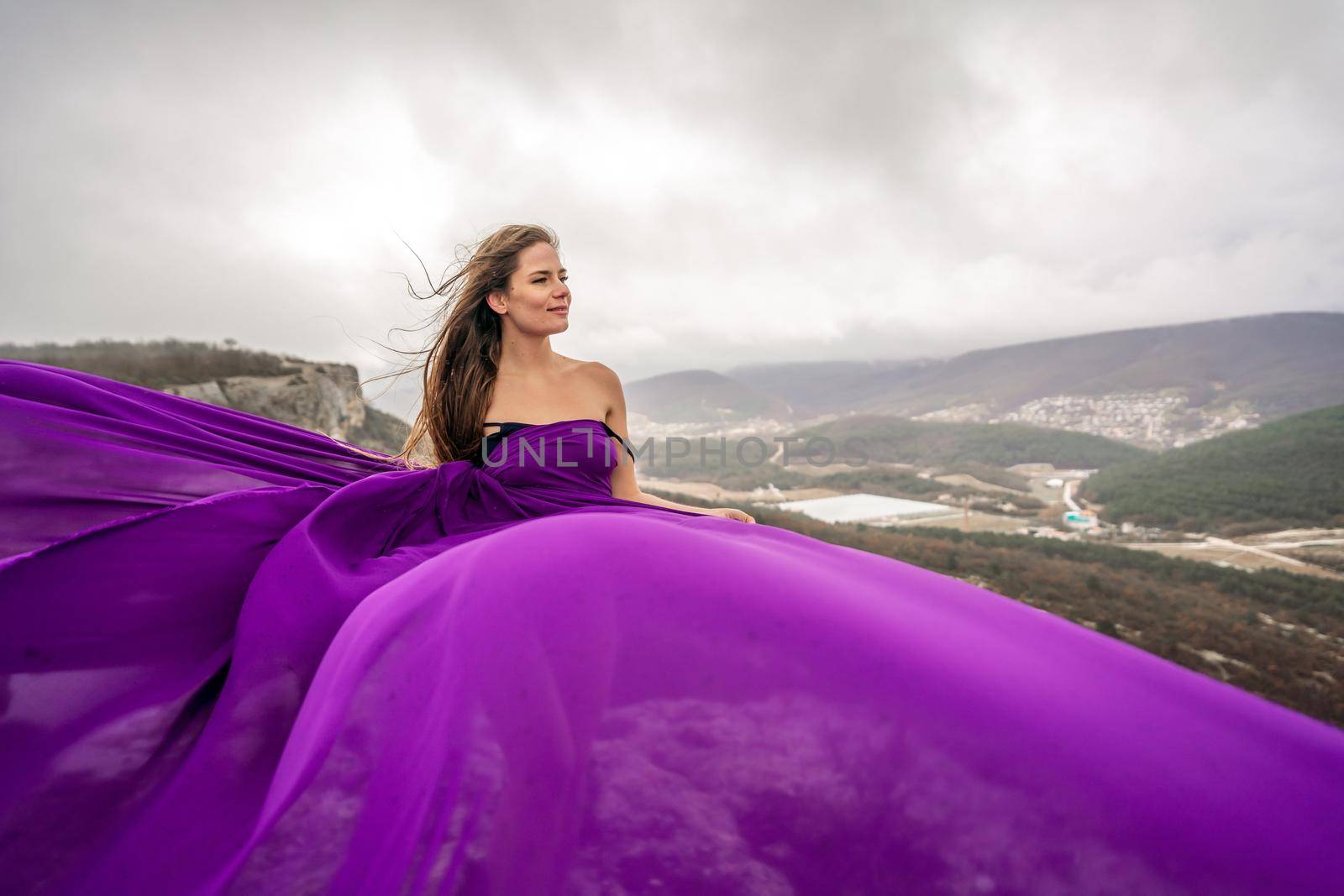 A woman with long hair is standing in a purple flowing dress with a flowing fabric. On the mountain against the background of the sky with clouds