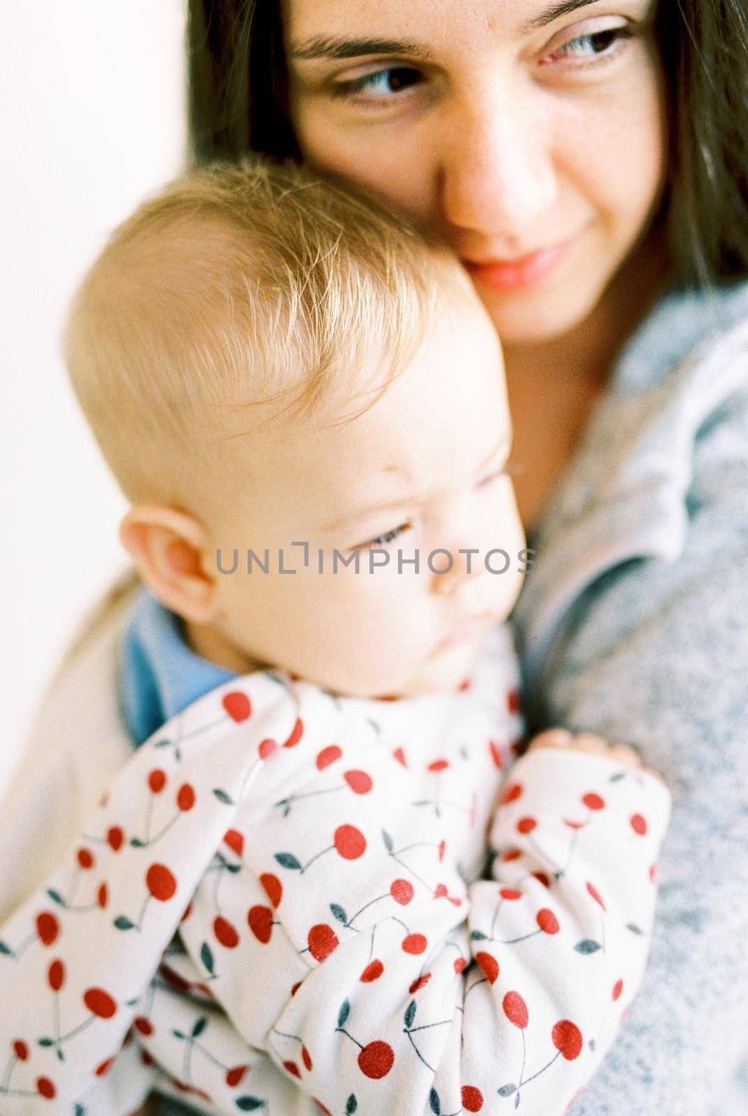 Mom hugs the baby to her and looks away. Portrait by Nadtochiy