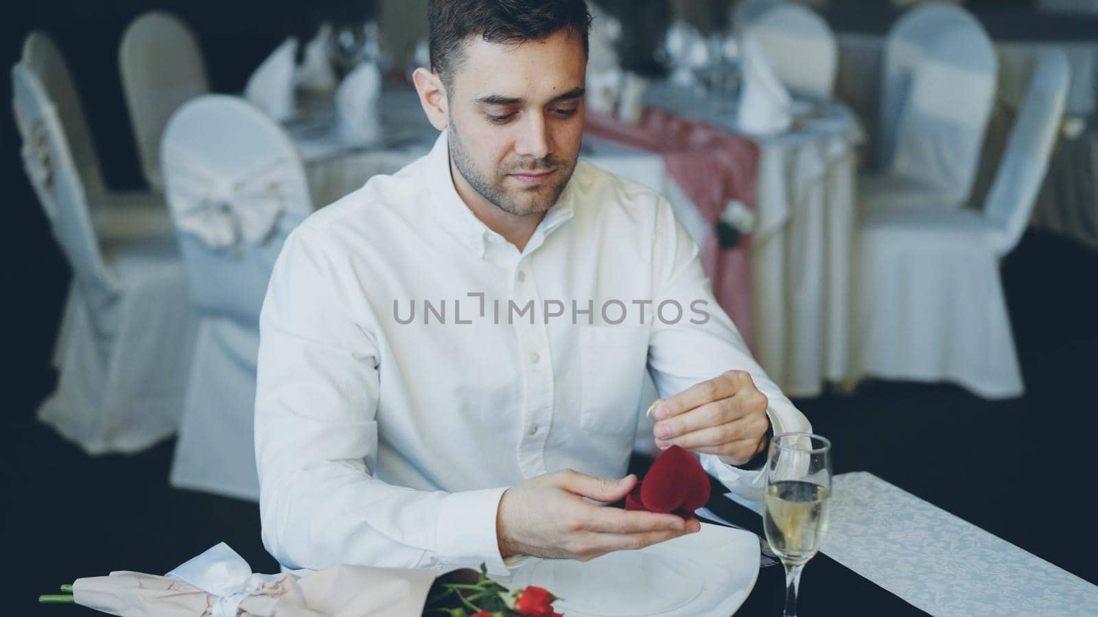 Loving handsome boyfriend is waiting for his girl in restaurant opening jewelry box and looking at ring. Bouquet of roses and glass of champagne are visible by silverkblack