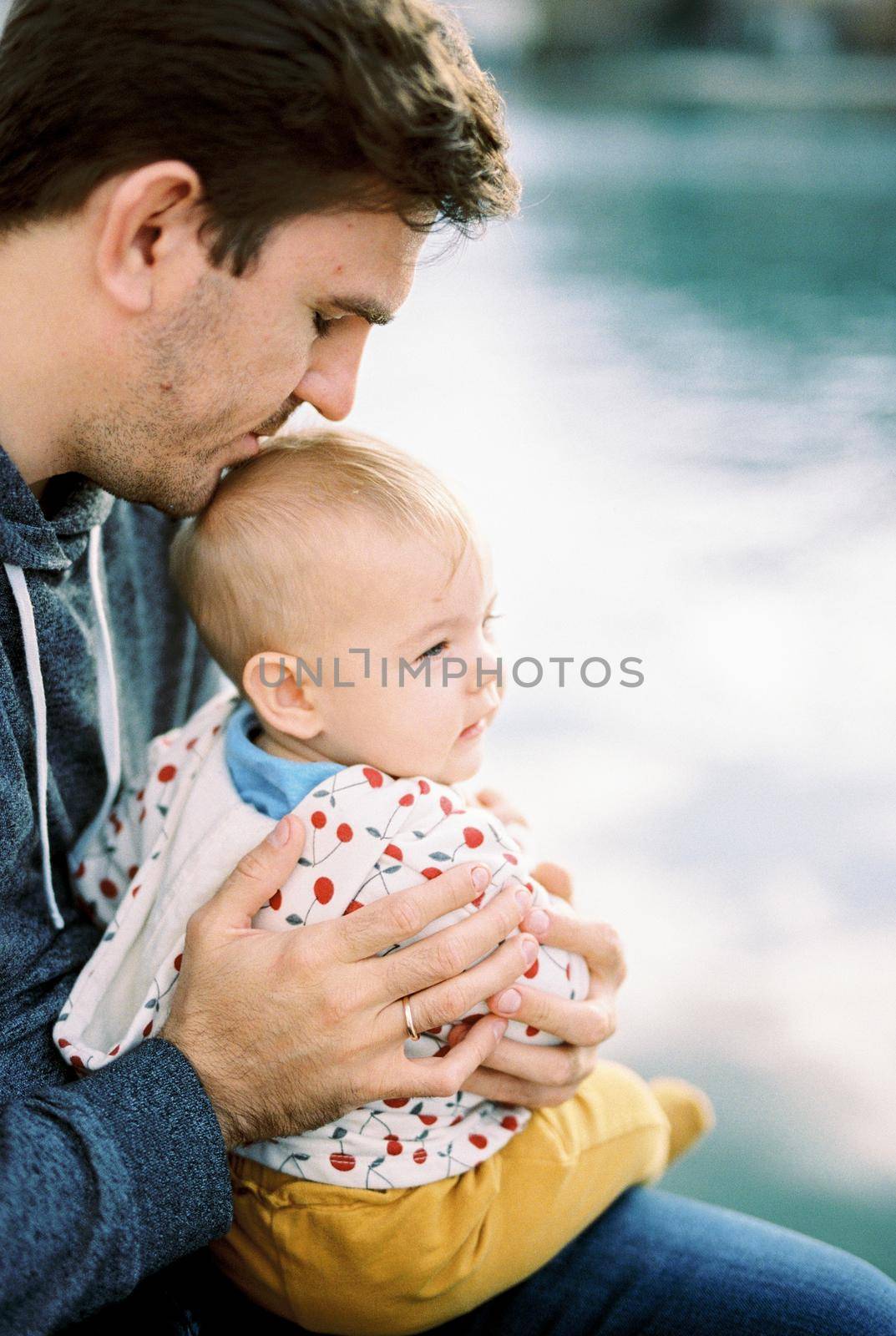 Dad hugs a baby sitting on his lap and kisses him on the top of his head by Nadtochiy