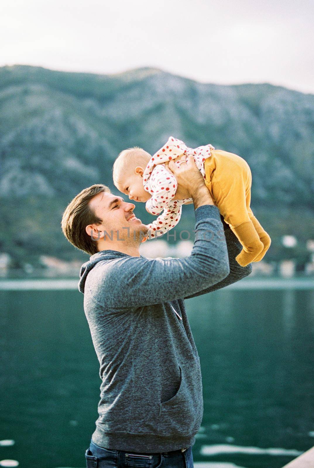 Smiling dad raises baby above his head against the background of water by Nadtochiy