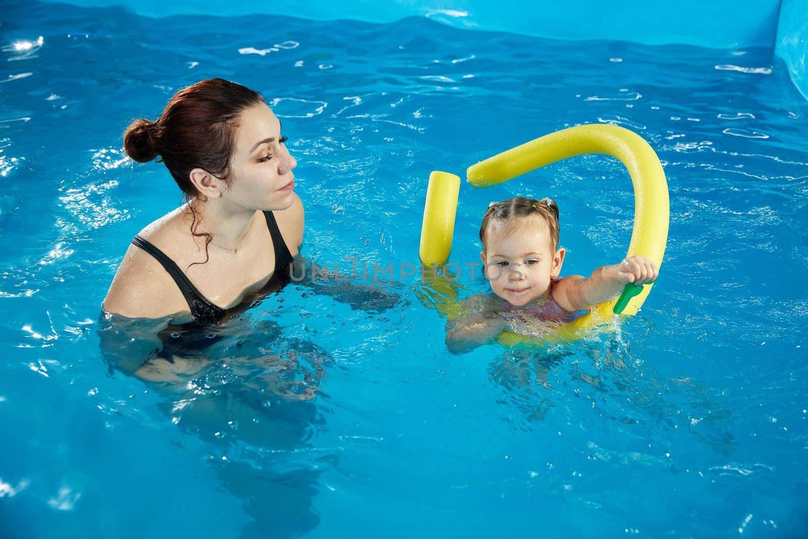 Preschool girllearning to swim in pool with foam noodle with young trainer by Mariakray