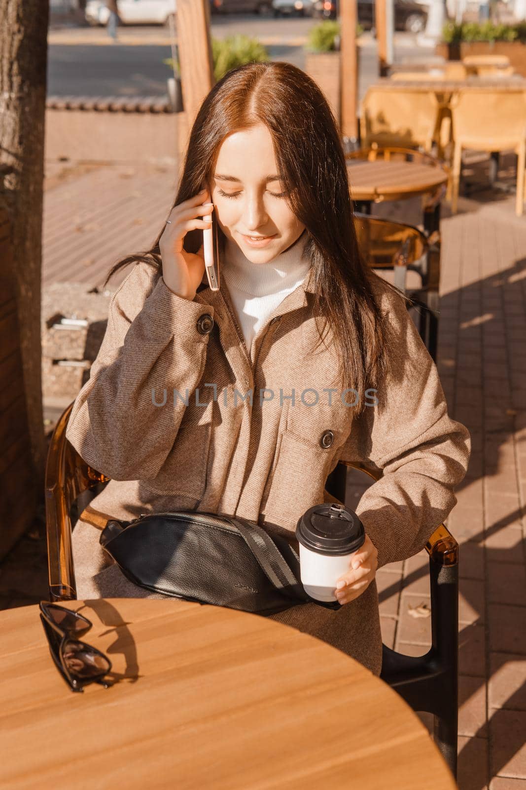 A stylish brunette woman is sitting at a table in an outdoor cafe with coffee and a phone. Autumn walk.