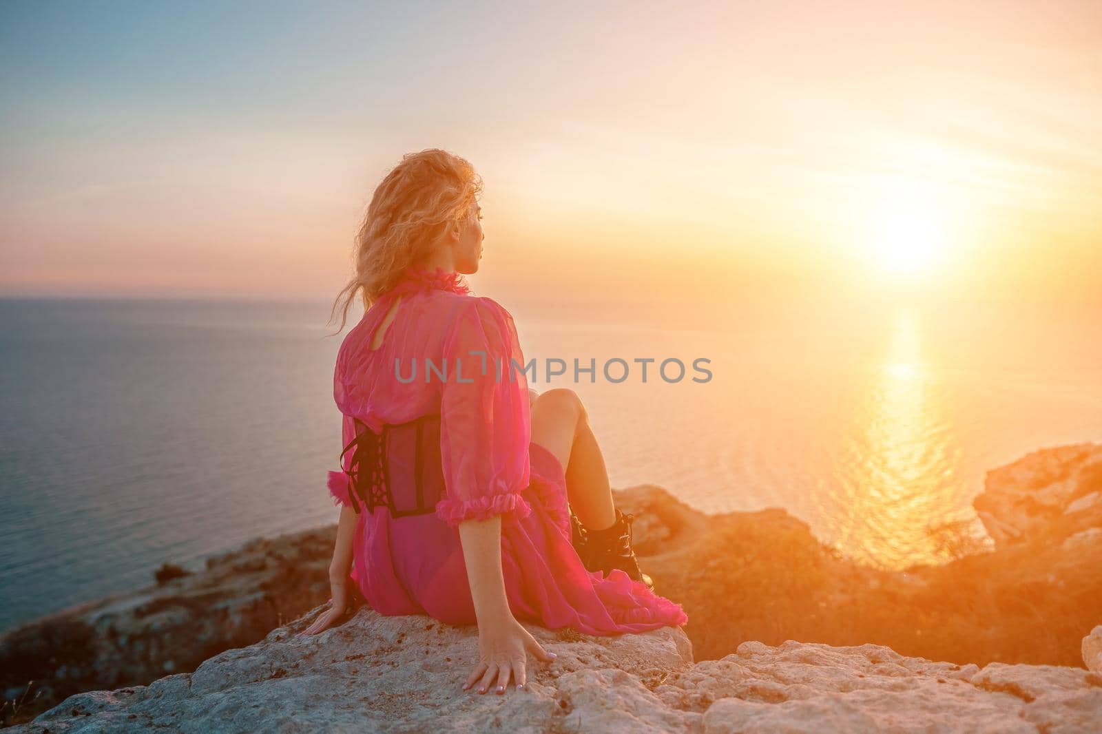 Side view a Young beautiful sensual woman in a red long dress posing on a volcanic rock high above the sea during sunset. Girl on the nature on blue sky background. Fashion photo