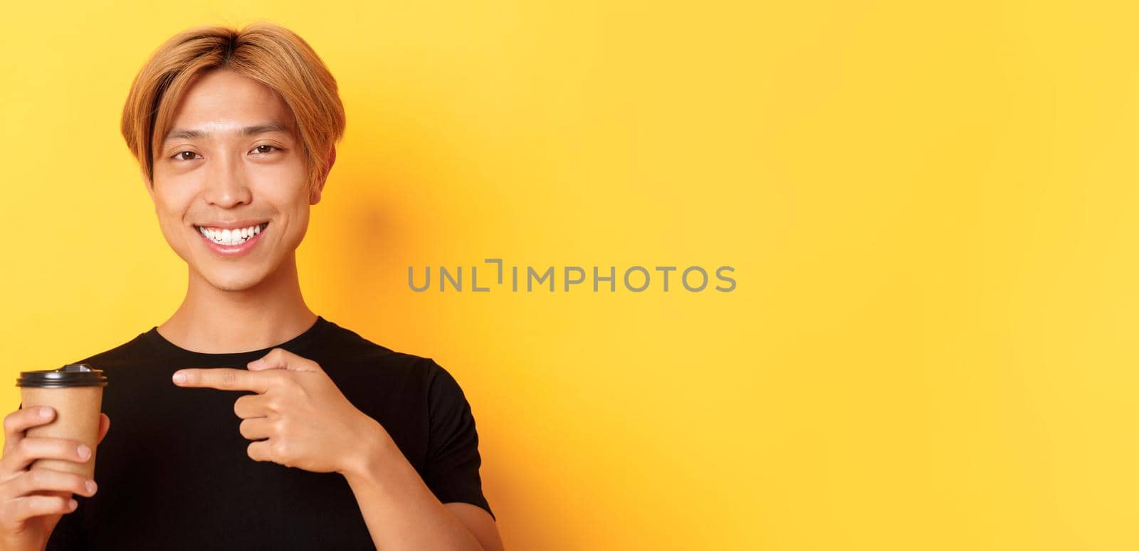 Close-up of satisfied handsome asian blond guy, smiling delighted and pointing finger at delicious cup of coffee, standing over yellow background.