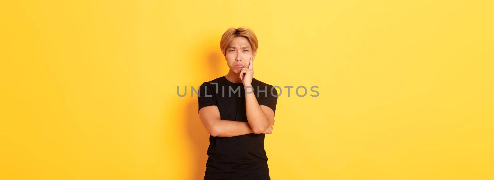 Upset thoughtful asian guy sulking and looking disappointed after thinking, standing yellow background by Benzoix