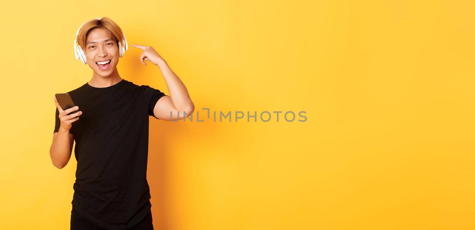 Happy satisfied asian guy likes music or podcast, pointing finger at headphones, holding smartphone, standing yellow background by Benzoix