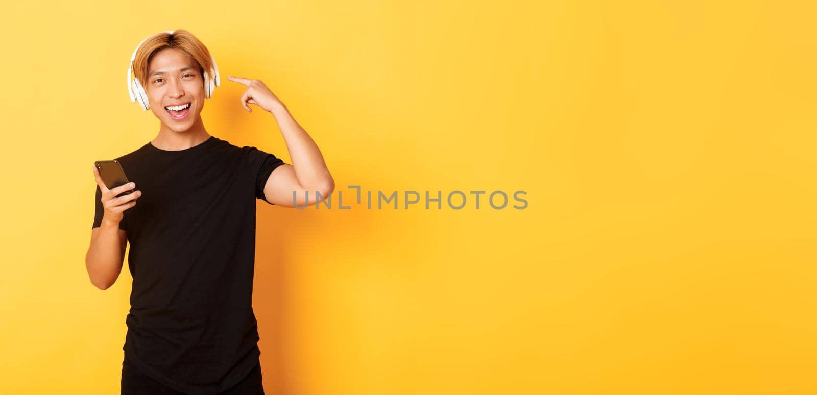 Happy satisfied asian guy likes music or podcast, pointing finger at headphones, holding smartphone, standing yellow background.