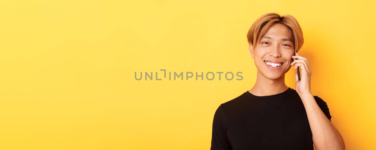 Close-up of handsome stylish korean guy smiling and talking on mobile phone, standing over yellow background.