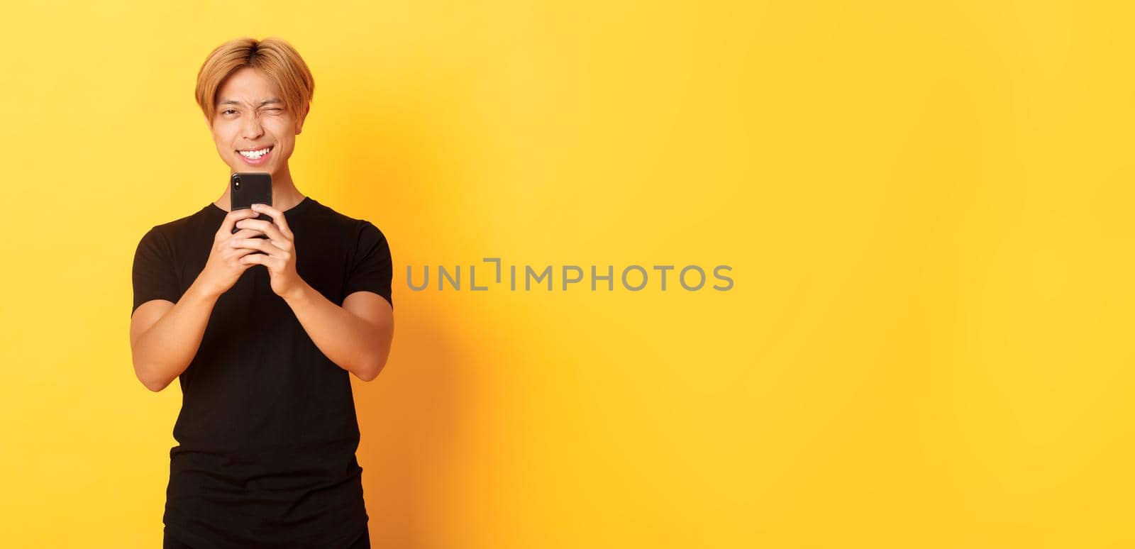 Stylish handsome asian guy taking picture on smartphone and smiling, photographing with mobile phone, standing yellow background by Benzoix
