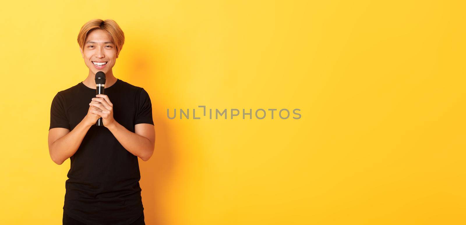 Portrait of charismatic smiling asian man holding microphone and singing karaoke, performing song, standing yellow background.