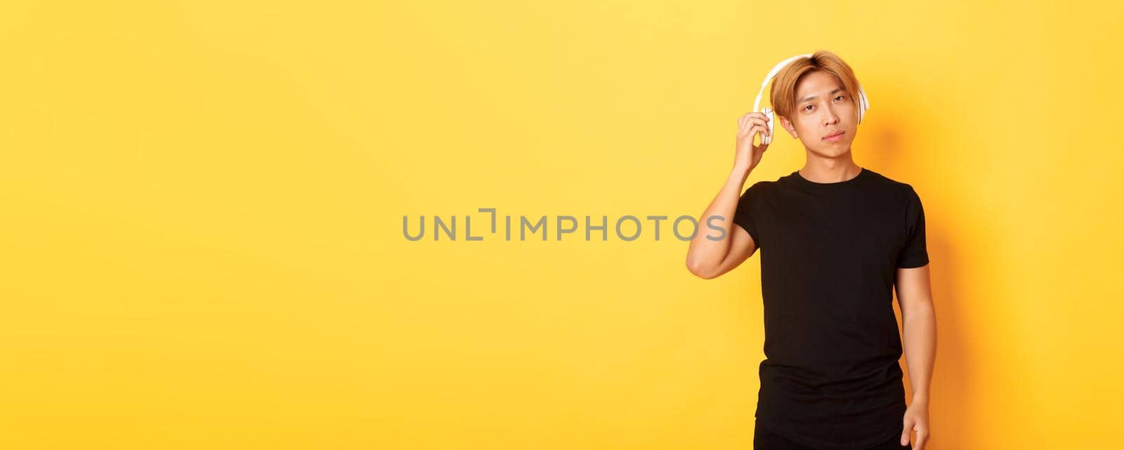 Young handsome asian guy with blond hair, take-off headphones to listen you, standing yellow background.