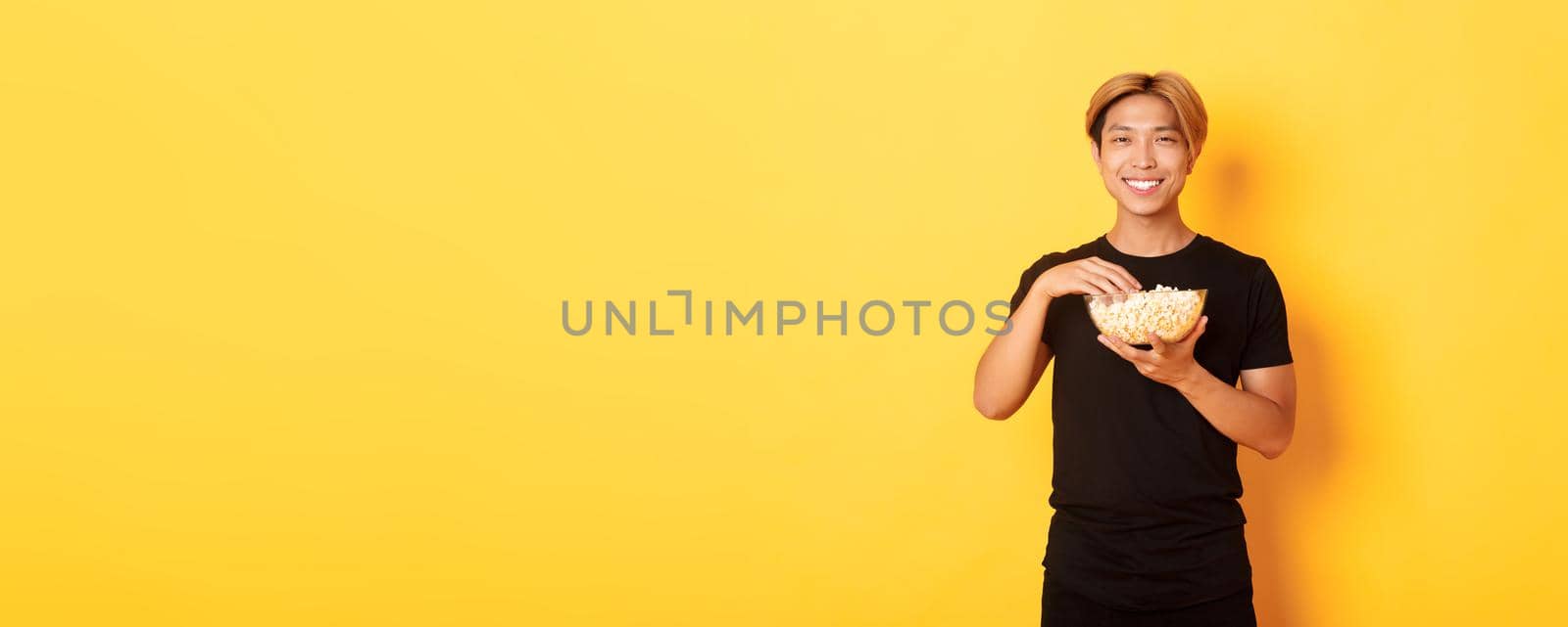 Pleased handsome korean guy smiling happy as enjoying watching movie or tv series, eating popcorn, standing yellow background.