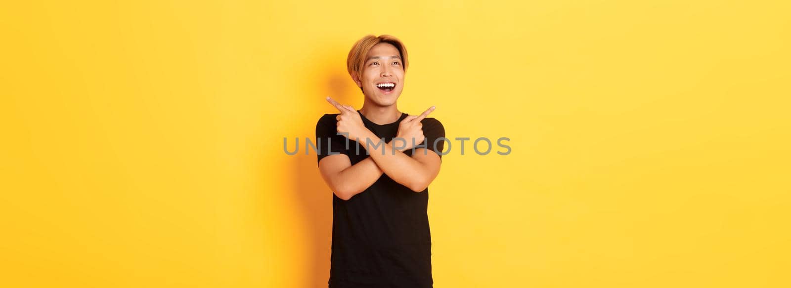 Portrait of happy smiling asian man with blond hair, looking amused left, pointing sideways at two variants, yellow background by Benzoix
