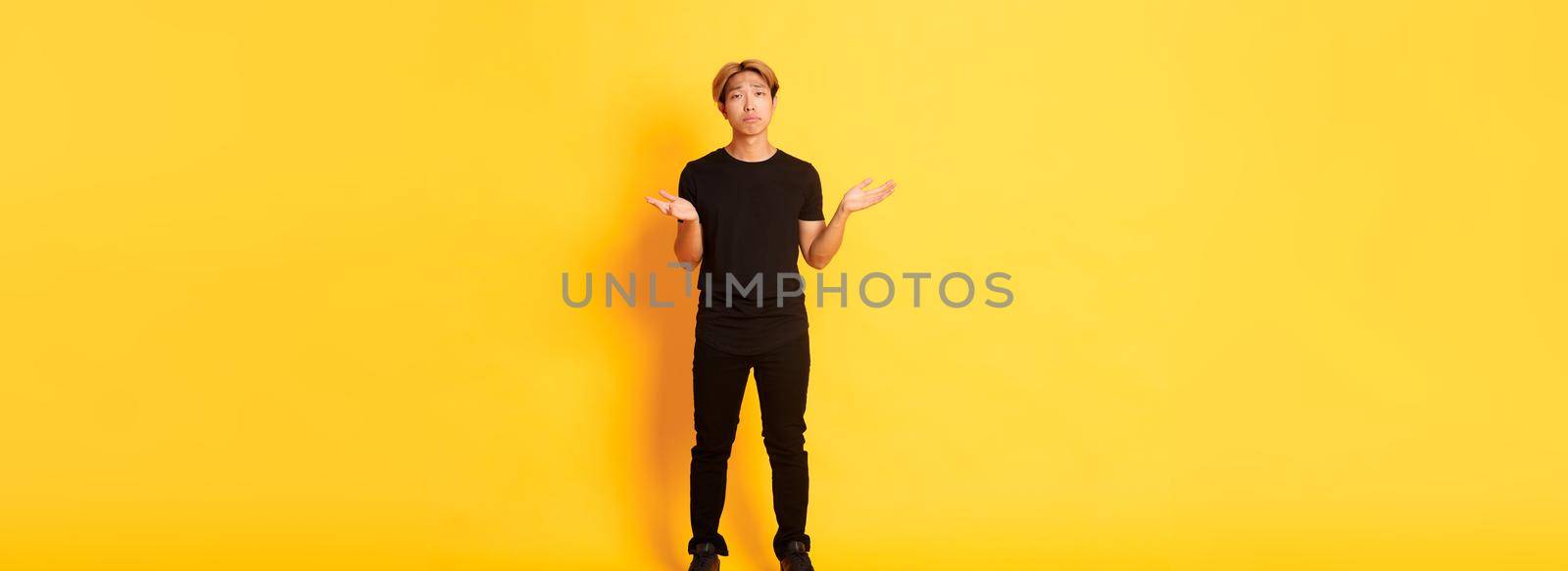Full-length of disappointed and frustrated asian guy shrugging, looking sad, yellow background.