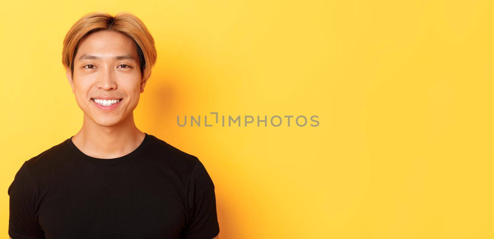 Close-up of handsome blond asian guy in black t-shirt, smiling happy at camera, standing over yellow background.
