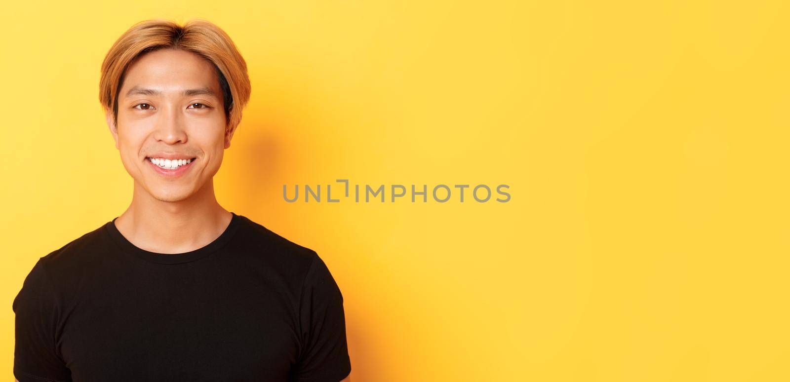 Close-up of handsome blond asian guy in black t-shirt, smiling happy at camera, standing over yellow background by Benzoix