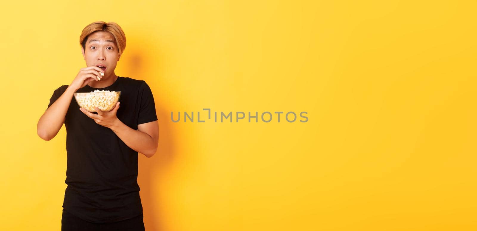 Excited and amazed asian guy watching interesting movie and eating popcorn from bowl, standing yellow background.