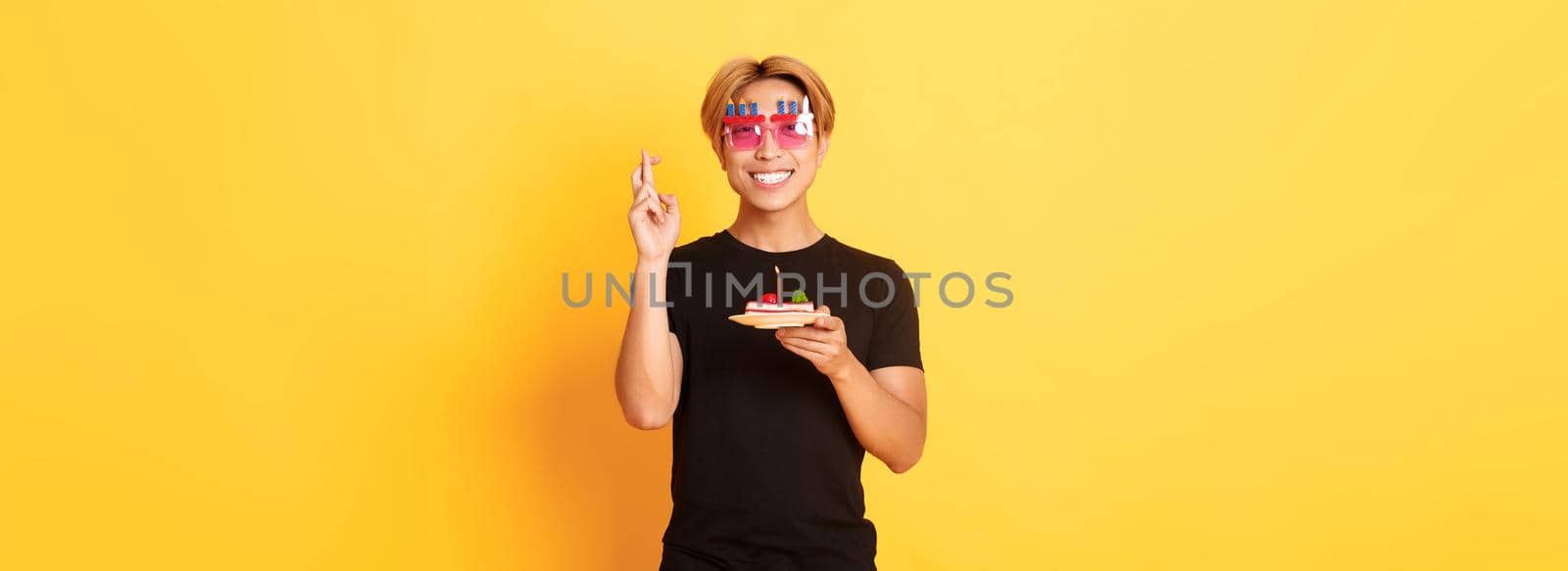 Portrait of hopeful attractive asian birthday guy, celebrating in funny party glasses, holding cake and fingers crossed while making b-day wish, yellow background.