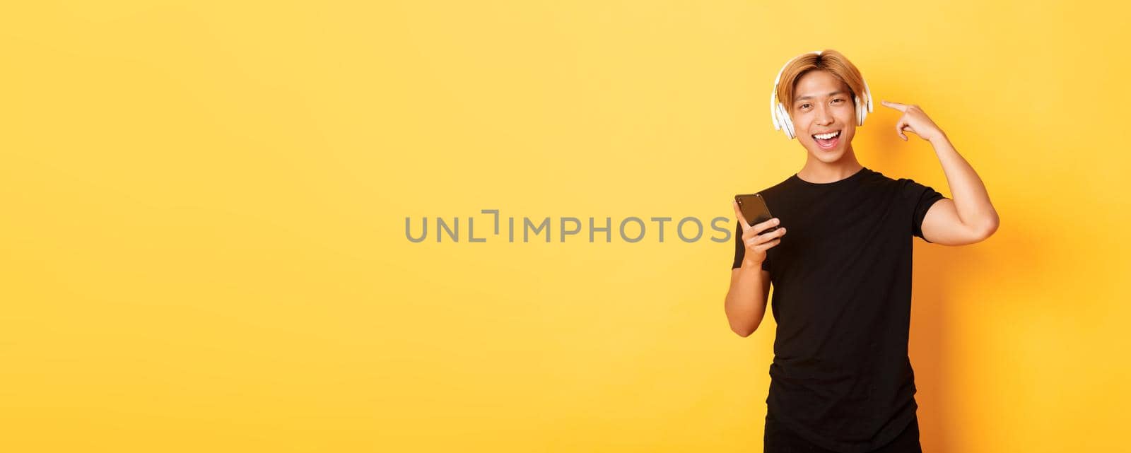 Happy satisfied asian guy likes music or podcast, pointing finger at headphones, holding smartphone, standing yellow background.