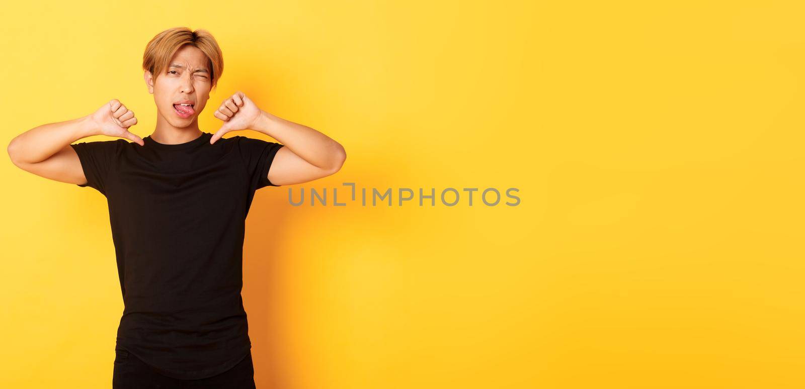 Portrait of sassy asian handsome guy showing thumbs-down and stick tongue disappointed, judge something bad, yellow background.