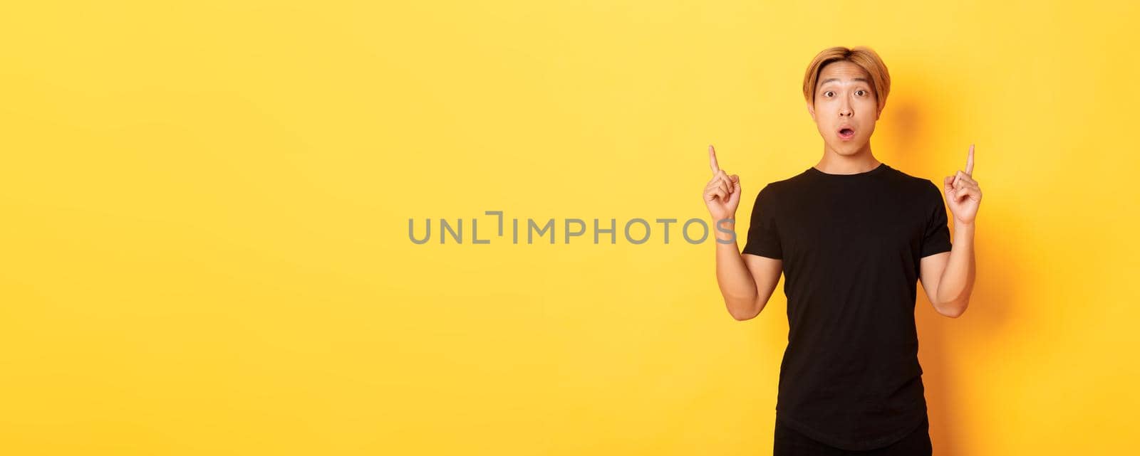 Portrait of astonished asian guy in black t-shirt, open mouth fascinated, pointing fingers up, yellow background by Benzoix